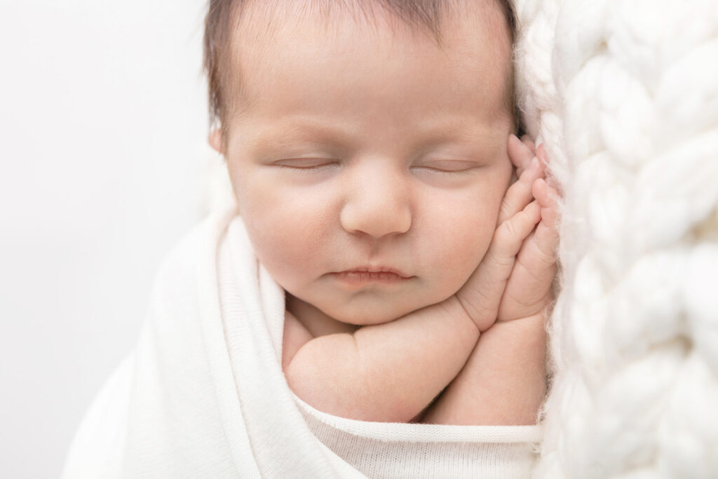 A newborn baby girl is photographed in shades of white and ivory, with her folded hands underneath her cheek. She sleeps softly on a chunky knit blanket