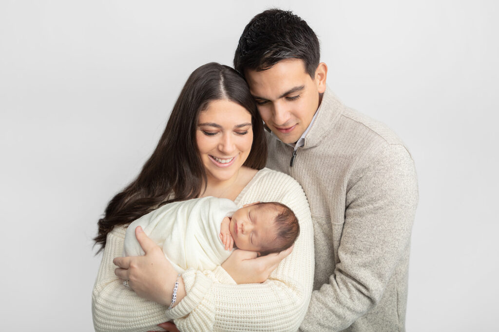 A new family of 3 is photographed by Karen Kahn in the Looking Up Photography studio in Greenwich, Connecticut. Mom and dad smile down gently at their newborn baby boy