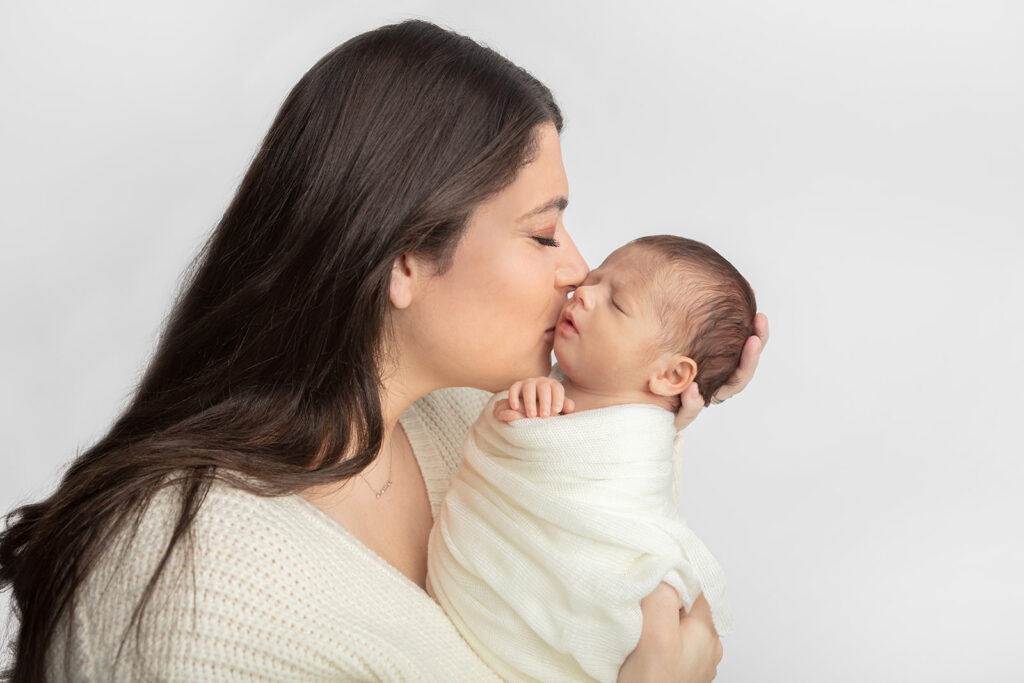 A newborn baby boy is photographed being held by his mom, who lifts him up to her face so she can kiss him. The pair match in different ivory textures, and are photographed against a white background