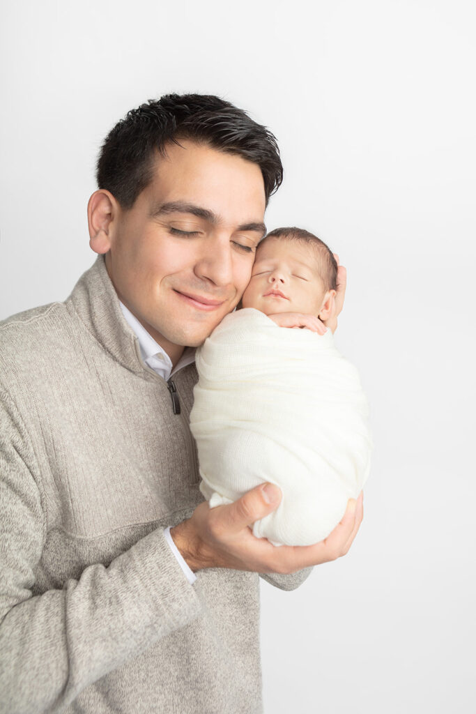 A newborn baby boy is photographed by Karen Kahn, in the Looking Up Photography studio. Matteo's dad holds him up so the pair are photographed cheek to cheek.