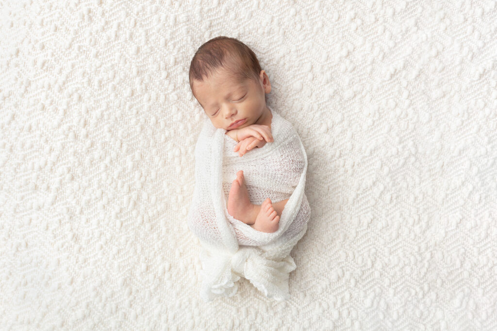Newborn baby Matteo lies with his hands and feet folded, lightly swaddled in an open knit swaddle, on a creamy textured blanket in the Looking Up Photography Studio