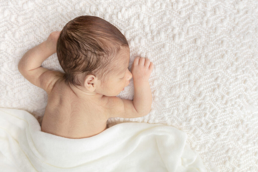 Newborn baby Matteo, with his fuzzy arms and back, and head full of hair, sleeps on his tummy with his mouth slightly open, looking very relaxed, photographed by Karen Kahn