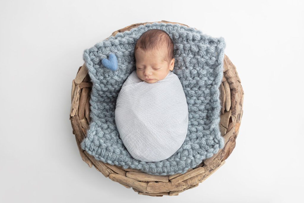 A farmhouse style newborn portrait, with newborn Matteo tightly swaddled in light blue, lies on a chunky knit, dusty blue blanket square in a driftwood basket, against a white backdrop