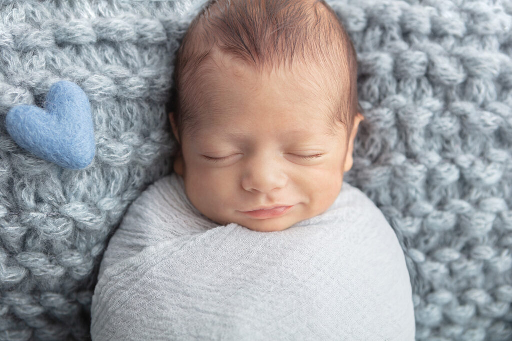 Smiling newborn baby boy appears to be smiling in his sleep. Baby Matteo is tightly swaddled in a gauzy, light blue swaddle, and a felted blue heart sits offset in the photo frame, giving the portrait a whimsical look.