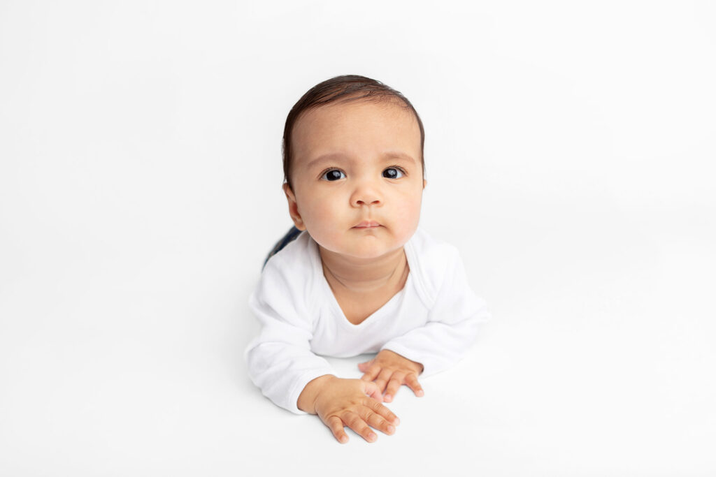 Baby Ezra is photographed in a simple white onesie, crawling toward the camera, against a white studio backdrop, in the Looking Up Photography studio