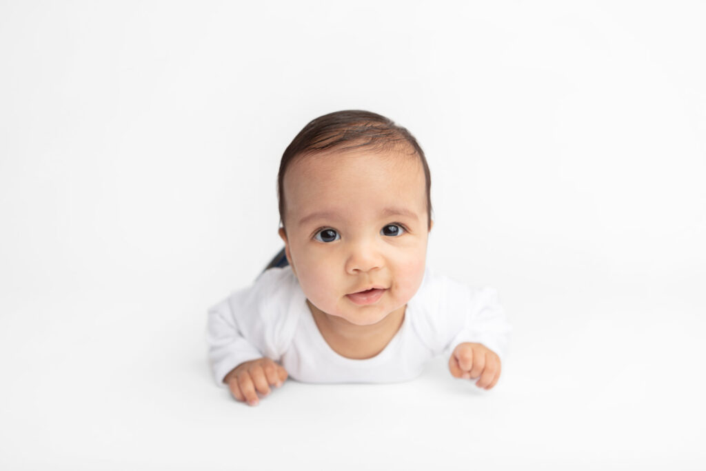 Baby Ezra is photographed with a slight smile or smirk on his face as he lifts his head and neck up to look at the camera during some studio tummy time