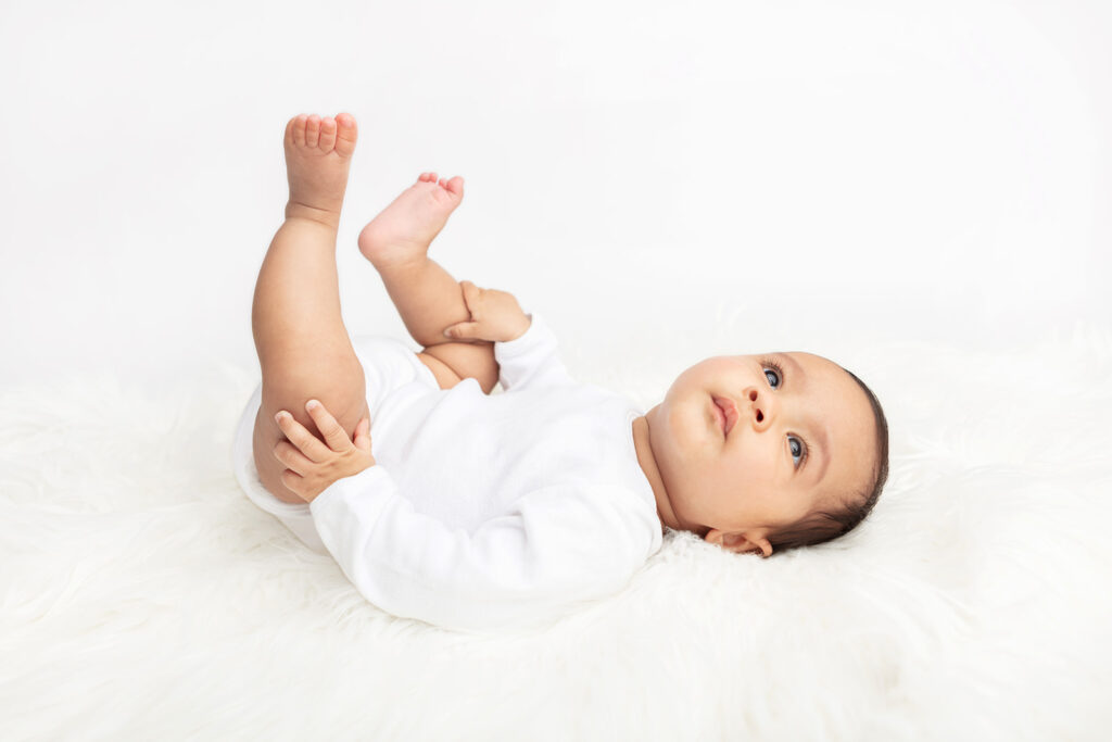 Baby Ezra lies on his back, holding his knees in his hands, looking pensively up at the ceiling. The baby is photographed wearing a simple white onesie