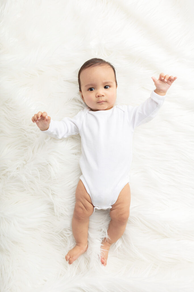 Baby Ezra is photographed wearing a simple white onesie, against the background of a creamy white flokati rug, his arms waving gently in the air