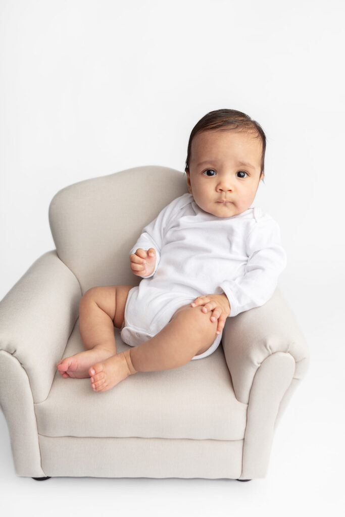 Baby Ezra is photographed for a milestone portrait session, sitting propped in a beige colored, overstuffed, baby sized cozy chair