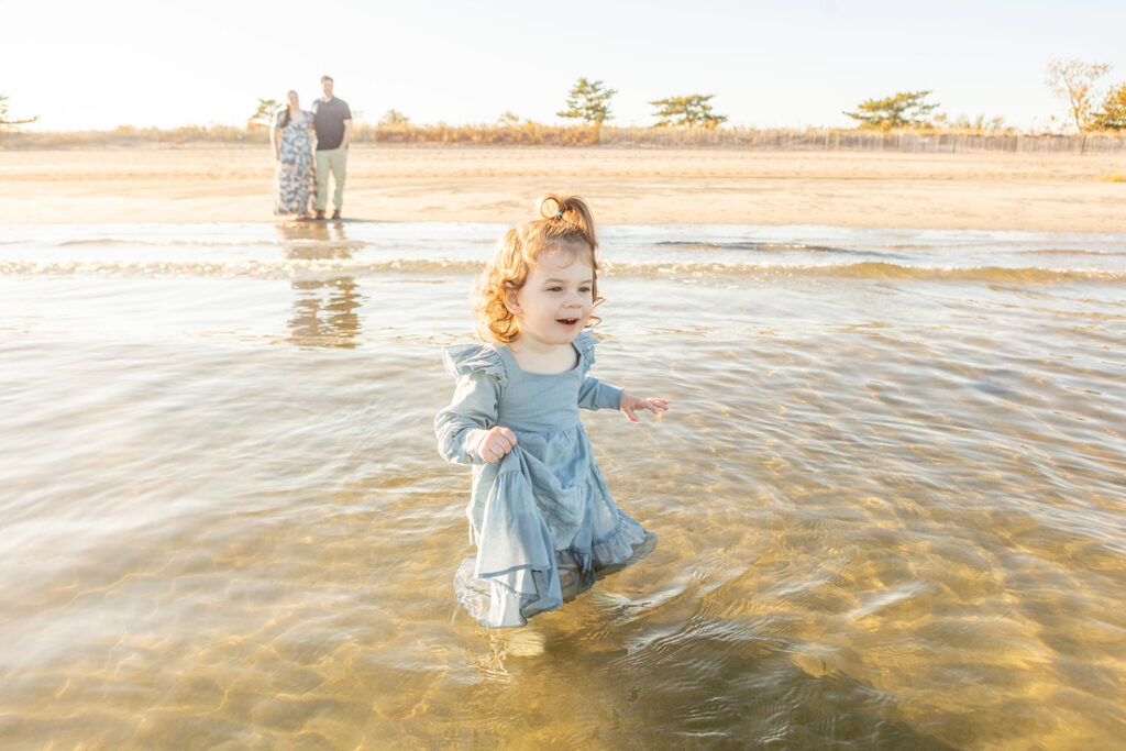 Two and a half year old Blake walks into the ocean for the very first time, delicately holding on to her denim-blue dress, her face filled with delight as her parents watch from the sand.