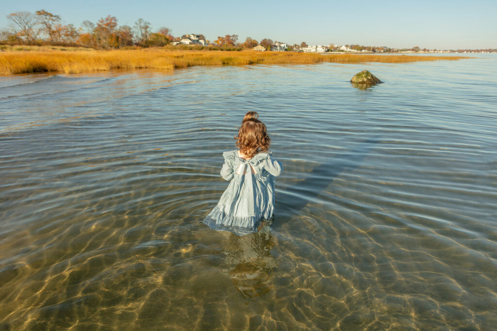 In a golden hour fall beach portrait, a little girl wades into the clear Atlantic ocean, her auburn curls highlighted by the golden light of the setting sun.