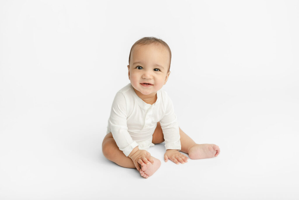A sweetly smiling baby Ezra is photographed sitting up for his milestone portrait session; he props himself up with his hands