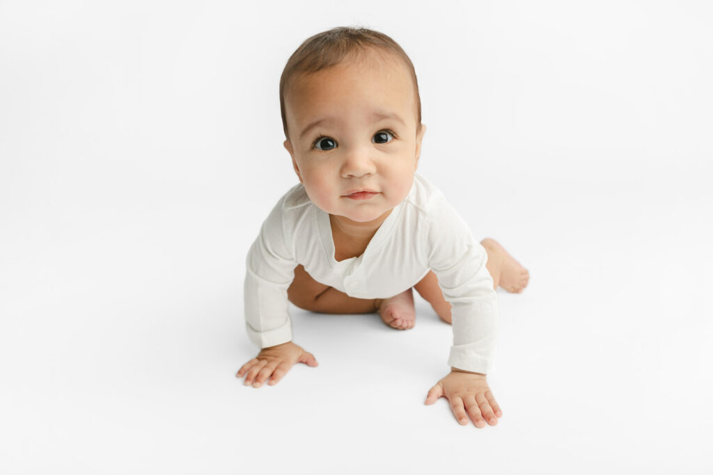 Baby Ezra is photographed in a white onesie, crawling toward the camera, in the Looking Up Photography studio in Greenwich, Connecticut