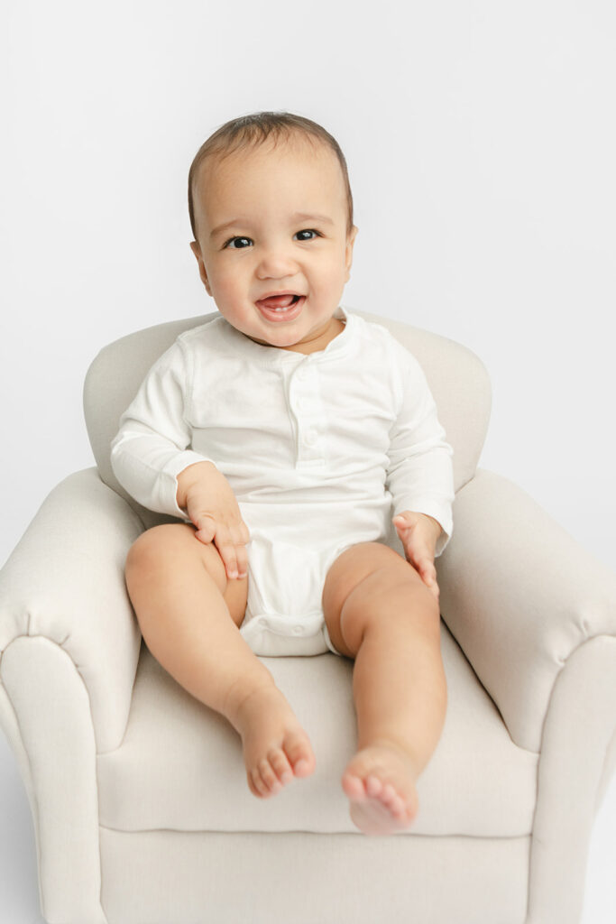 A smiling Baby Ezra sits independently in the baby-sized overstuffed chair in the Looking Up Photography studio in Greenwich, Connecticut