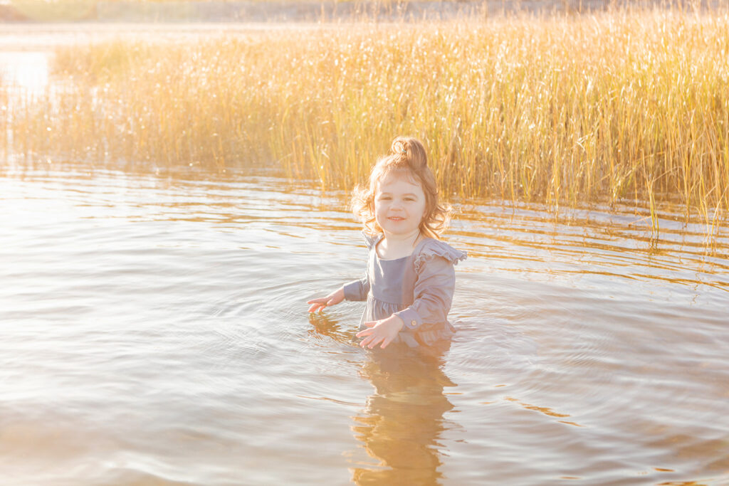 A two year old little girl is photographed during her first visit to the ocean, bathed in the hazy golden light of the setting sun, sea grasses behind her