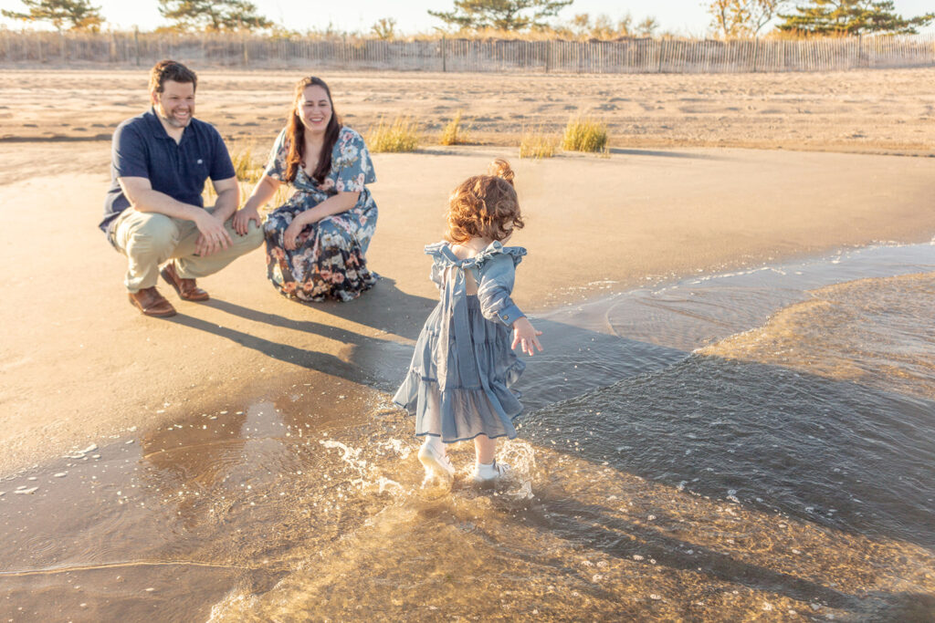 A duo of Connecticut parents squat on the beach, smiling at their two year old daughter as she splashes through the surf of a Connecticut beach at sunset