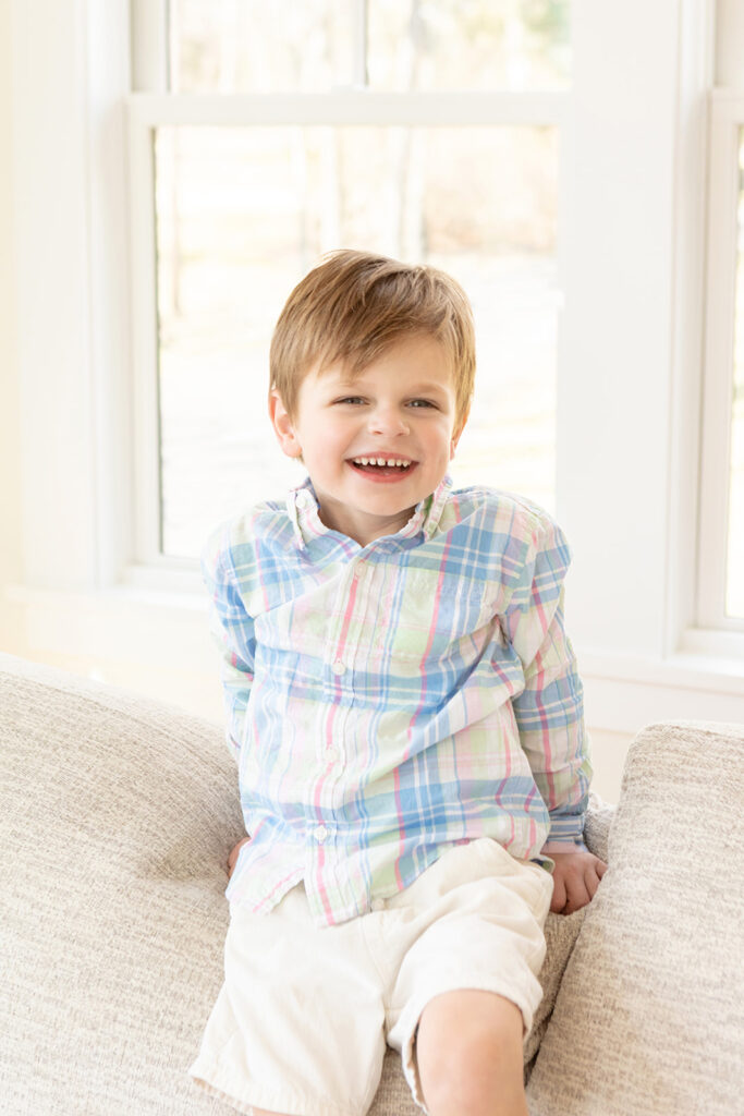 a 3-year-old boy wearing a classic New England style outfit of chino shorts and a pastel madras plaid button down shirt is photographed in his family's home