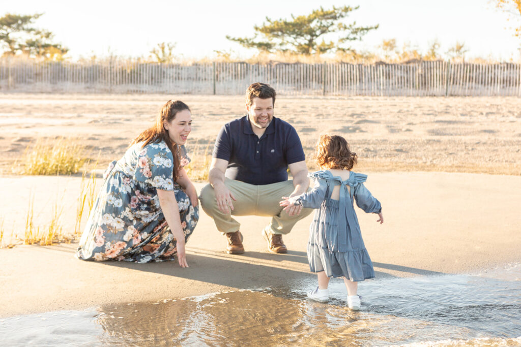 A mother and father laugh and smile a their two year old daughter as she experiences the ocean for the very first time. The family are photographed by Looking Up Photography
