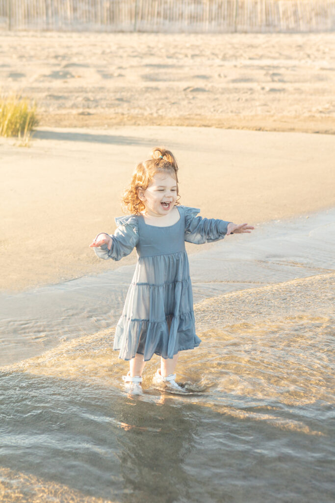 A toddler girl with strawberry blonde curls appears to be conducting the ocean as she laughs in the water at sunset. The portrait has a Moana quality to it!
