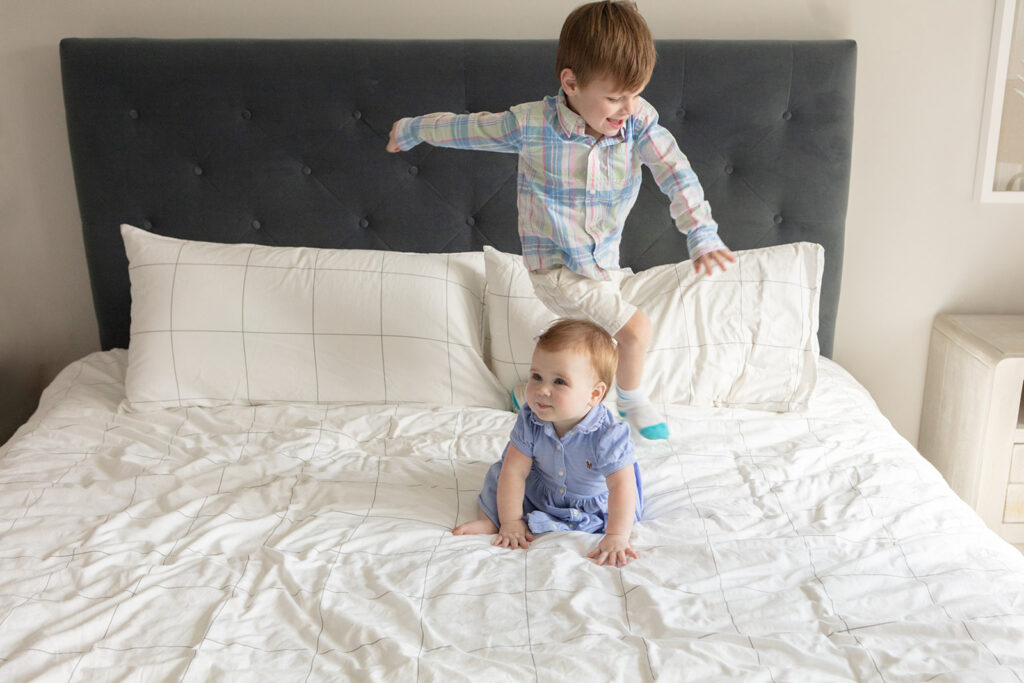 a 3-year-old boy and his 9-month-old little sister are photographed jumping on the styled bed of their parents' modern farmhouse style Connecticut home