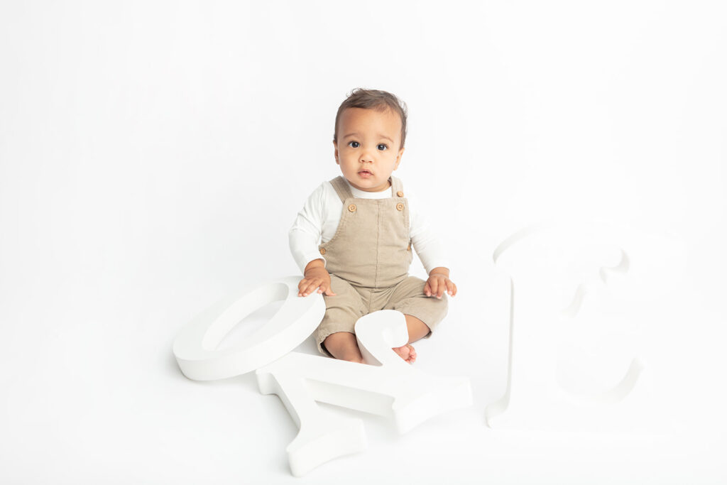 Baby Ezra is photographed for his first birthday in the Looking Up Photography studio, holding wooden white letters that spell out the word "ONE"