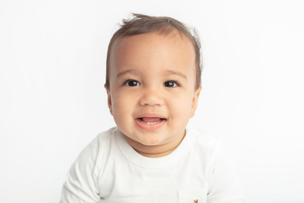 Baby Ezra looks just like a Gerber baby as he smiles at the camera showing off some new teeth on bottom, and his big, dark eyes; photographed against a white backdrop