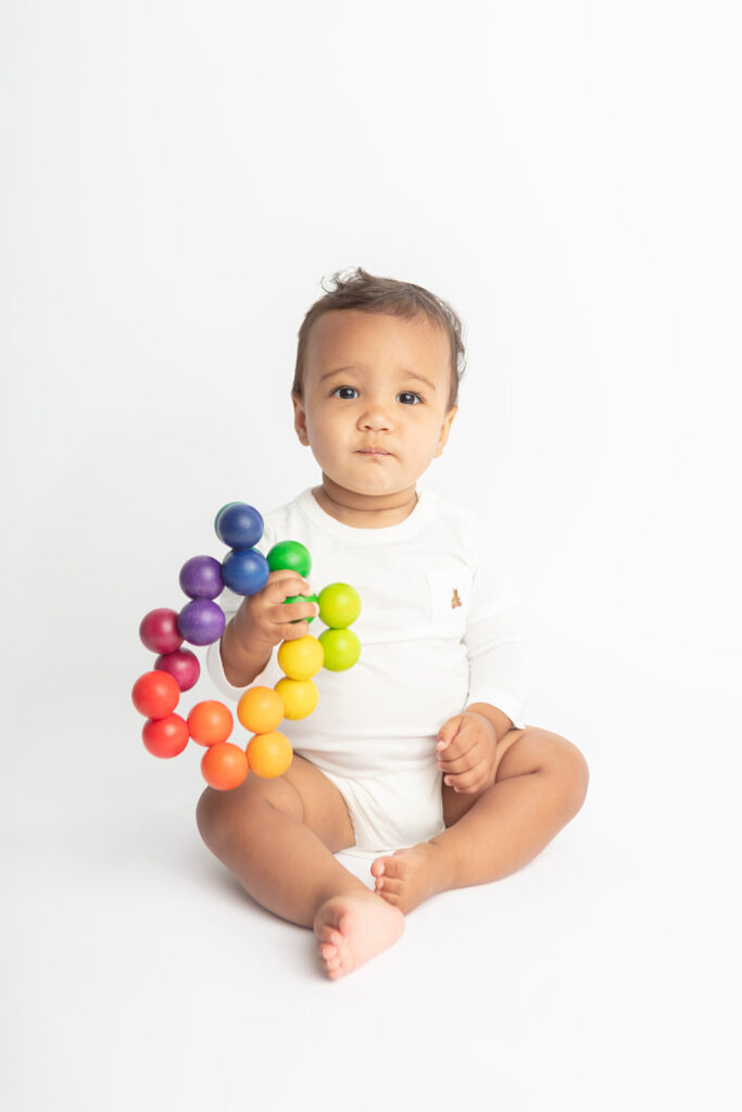 Baby Ezra is photographed in a white onesie with a small embroidered teddy bear on it, holding a rainbow ball toy in the Looking Up Photography studio