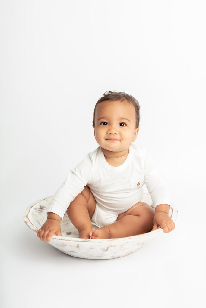 Baby Ezra puts a funny little smile on his face as he sits in a distressed, farmhouse style, white washbin, holding on to the sides of in the studio