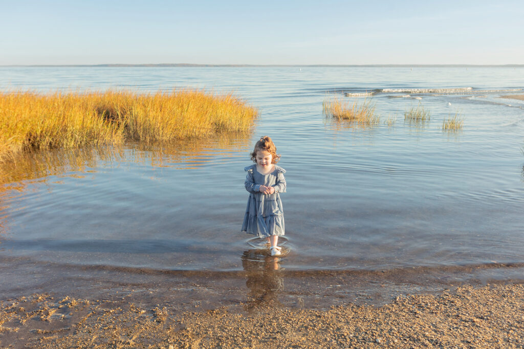 A little girl with strawberry blonde curls walks calmly out of the ocean as if she came from it herself. Gentle waves and seagrasses frame the portrait background