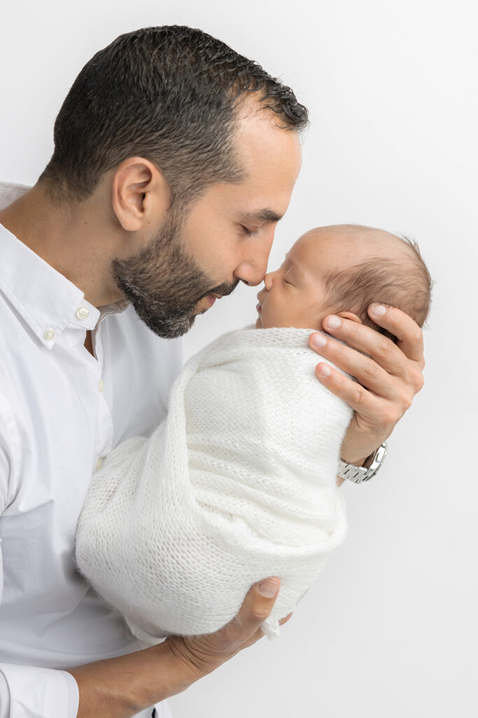 A new dad, wearing a silver watch and a white oxford shirt, closes his eyes and touches noses with his newborn baby son, who is sleeping in his hands, in the Looking Up Photography studio