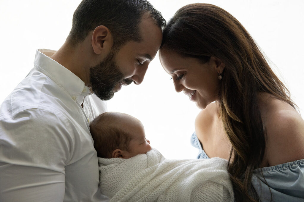A beautiful silhouette-style portrait of a dad, mom, and their newborn baby son. Mom and dad look down at their son, smiling, as he snoozes in between them