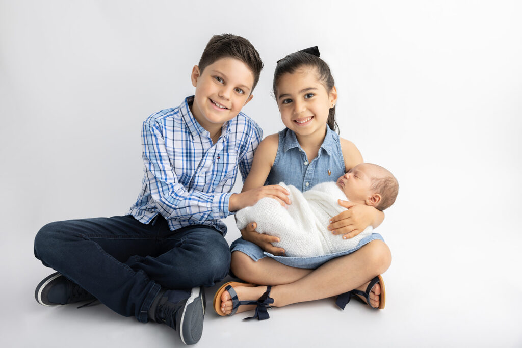 Proud big brother Luca and big sister Mila hold their newborn baby brother, Mateo, and smile at the camera as they sit criss cross apple sauce on the floor of the Looking Up Photography studio