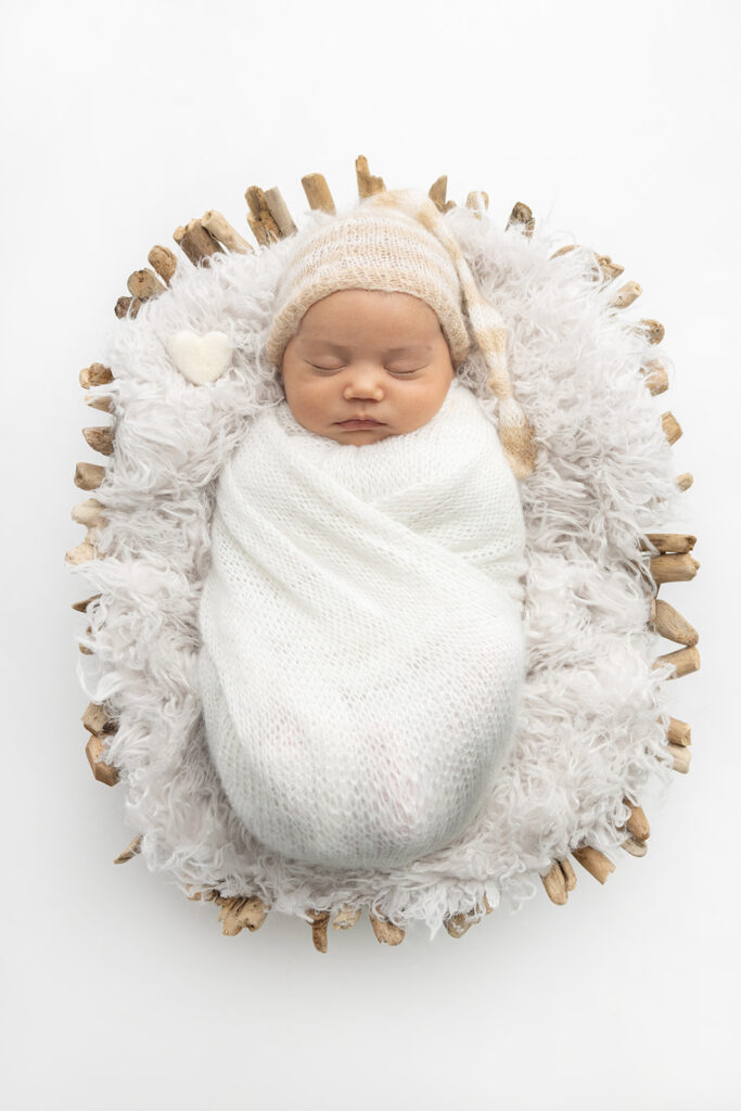 Newborn Mateo is photographed sleeping in a driftwood basket. He is wrapped in creamy white and wears a neutral striped stocking cap