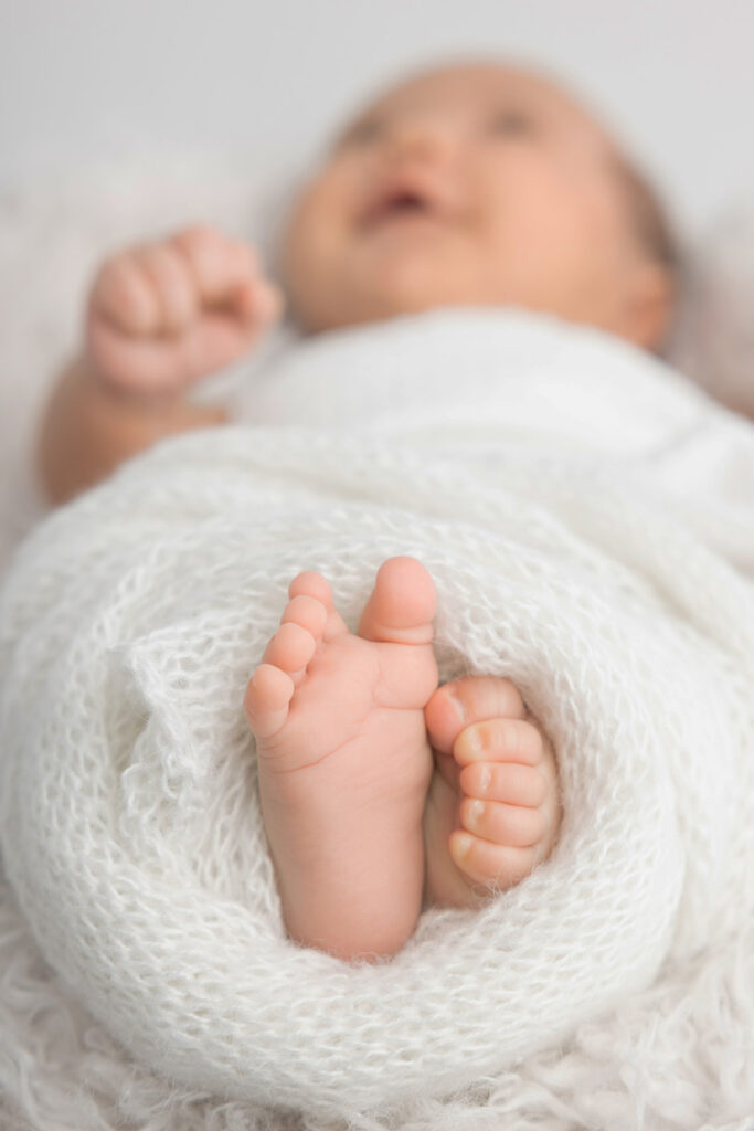 The sweetest portrait of newborn baby Mateo's feet and toes peeking out from a loose knit creamy white blanket. In the background, you can see that baby Mateo is happy and awake