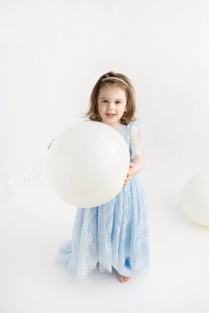 after a wardrobe change, three year-old Blake is photographed holding a large white balloon; she wears a lacy, light blue dress reminiscent of the Disney princess Cinderella