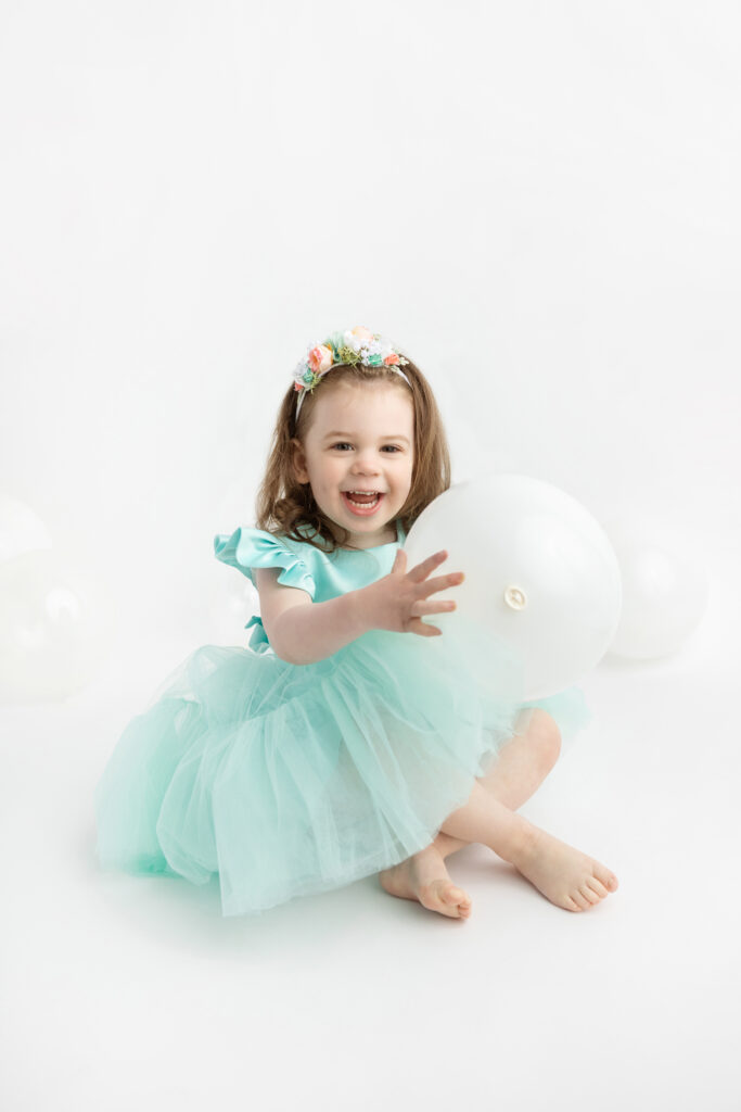 three year-old little girl Blake sits with her legs lightly crossed, in a sea foam green dress with a tulle skirt; she holds a white balloon and there are white and clear balloons in the background of the studio