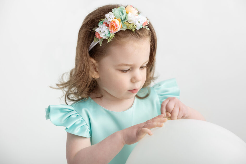 a three year-old little girl investigates a white balloon; she wears a sea foam green dress and a coral and sea foam green and white headband; the background is white