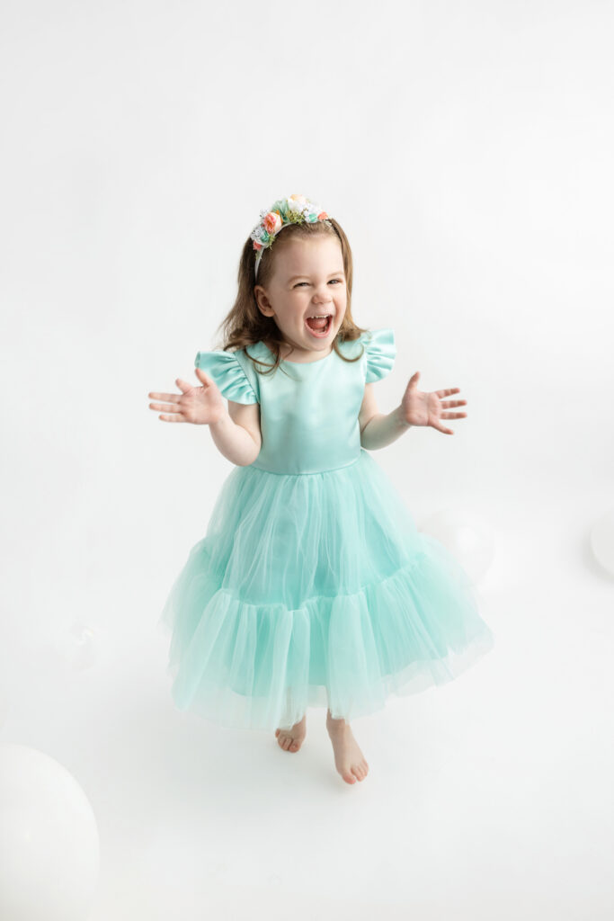 three year-old Blake is photographed for a milestone session in the Looking Up Photography studio; she has a big, delighted smile on her face, her hands out as if she's about to clap them together