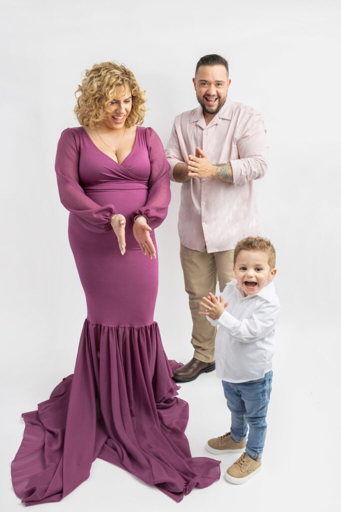 A young family of three plays a clapping game and dances to JLO, Daddy Yankee, and Lady Gaga, during their studio family maternity session captured by Karen Kahn.