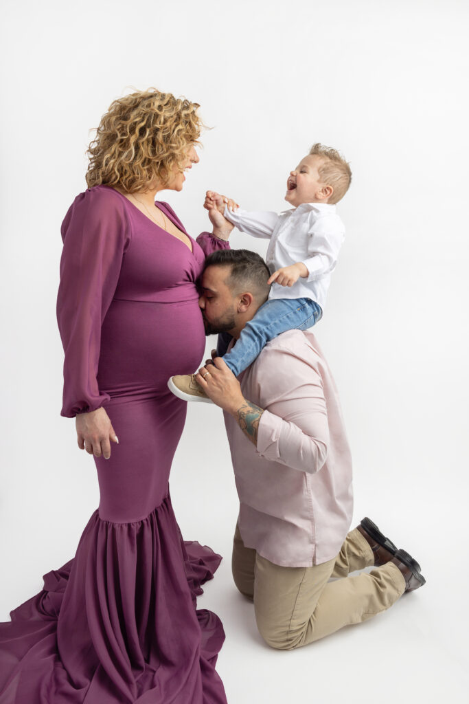 In a playful family maternity portrait, pregnant mama and toddler son laugh as the little boy sits on his dad's shoulders. Dad is kneeling in front of his wife, kissing her pregnant belly.