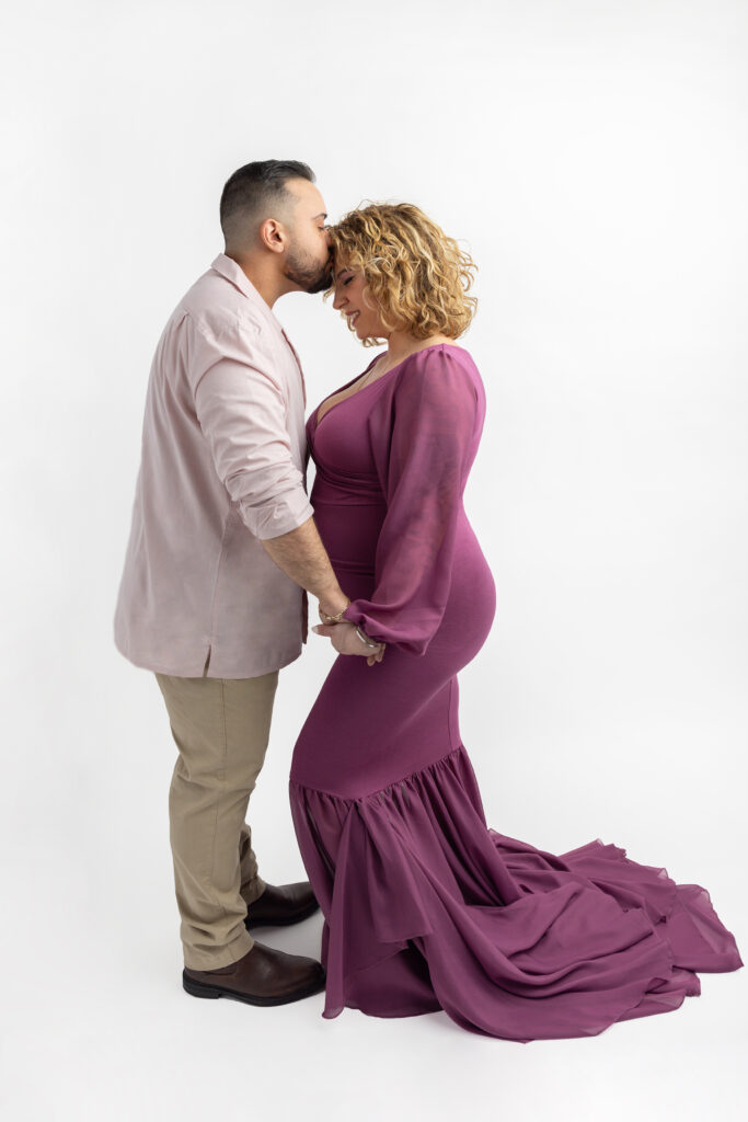 A proud papa to be in a pink and khaki linen suit, kisses his wife on the forehead. She smiles and closes her eyes as the pair hold hands, photographed during their studio maternity session in Connecticut.