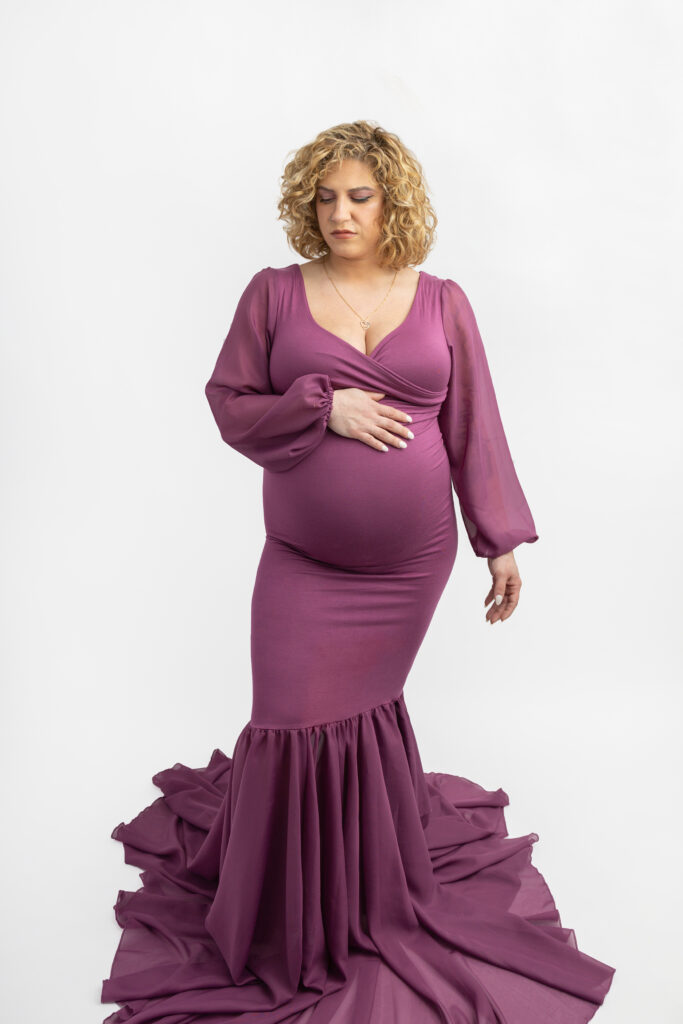A mother to be with short, very curly hair, pulls a pose during her studio maternity session with Karen Kahn, of Looking Up Photography. Courtney wears a maternity gown with sheer sleeves.