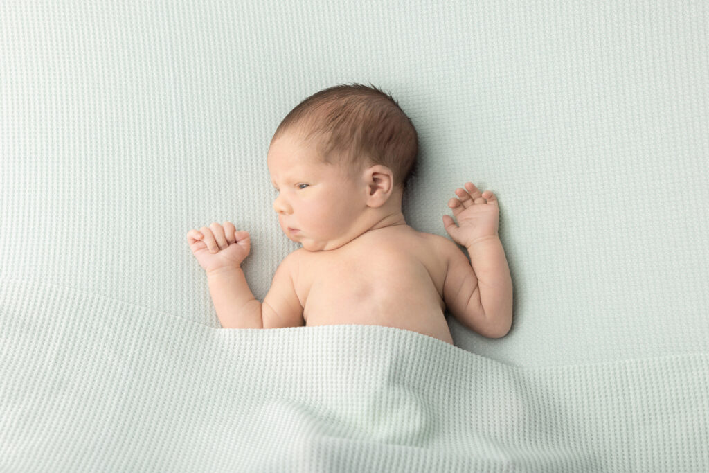 A newborn twin boy is pictured awake, lying on a waffle knit light green blanket, looking slightly concerned as he stares to the left of the photo, photographed in the Looking Up Photography studio.