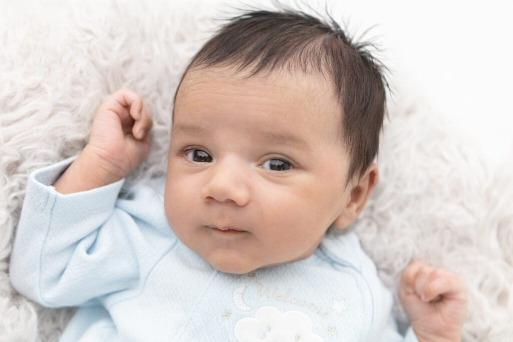 A newborn baby boy with dark eyes and hair looks at the camera inquisitively, with almost a little smirk on his sweet face. He wears a baby blue onesie with embroidered moon, stars, and a cloud.