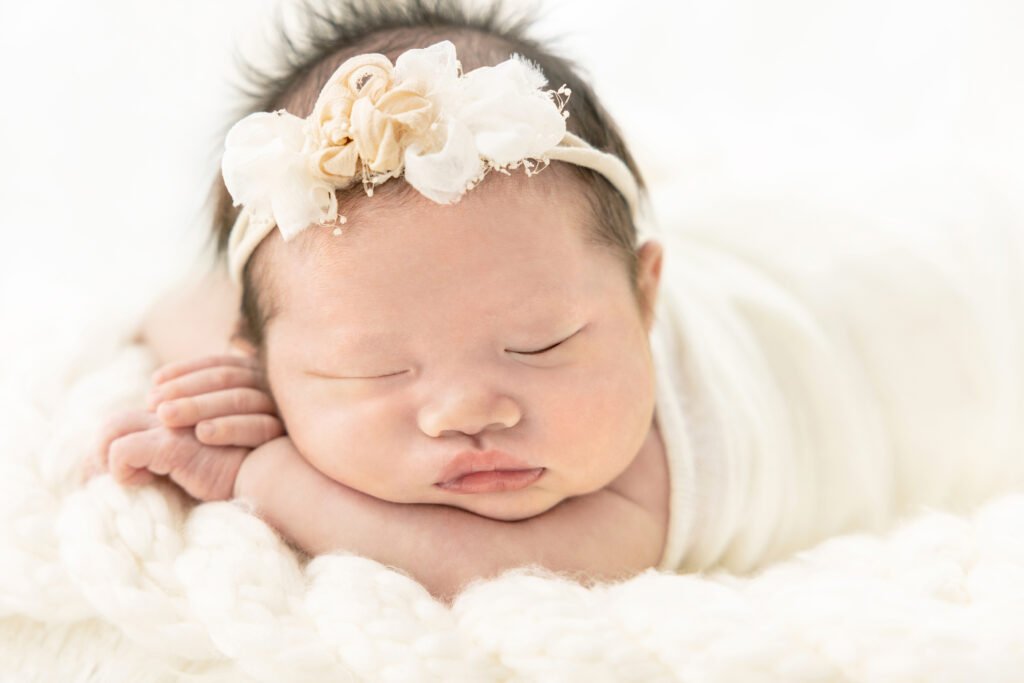 newborn baby Chloe sleeps on her folded arms, her face resting in as relaxed of a state as a newborn baby can be! she wears a white and ivory stretchy headband