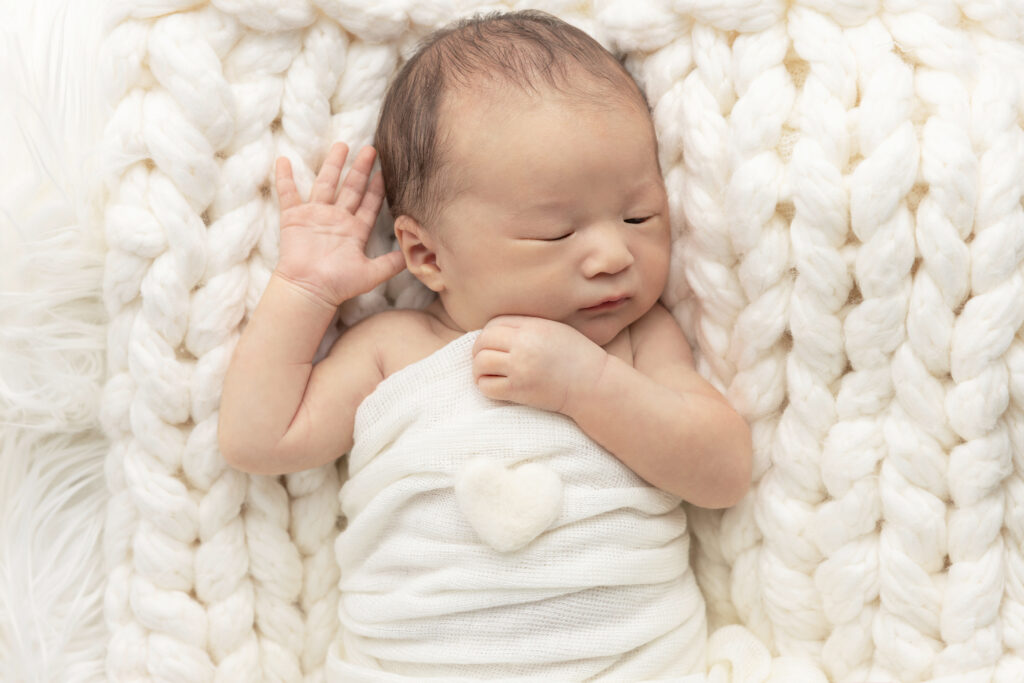 a newborn baby boy is photographed lying on a chunky, ivory knit blanket, lightly swaddled under his arms; a felted heart in an off white color lays on his chest