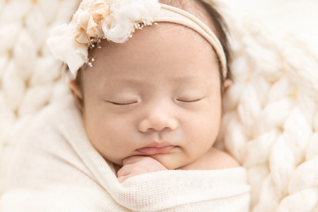 a sweet, newborn baby girl named Chloe, sleeps peacefully with her little hand emerging from her loosely wrapped swaddle, enveloped in shades of ivory