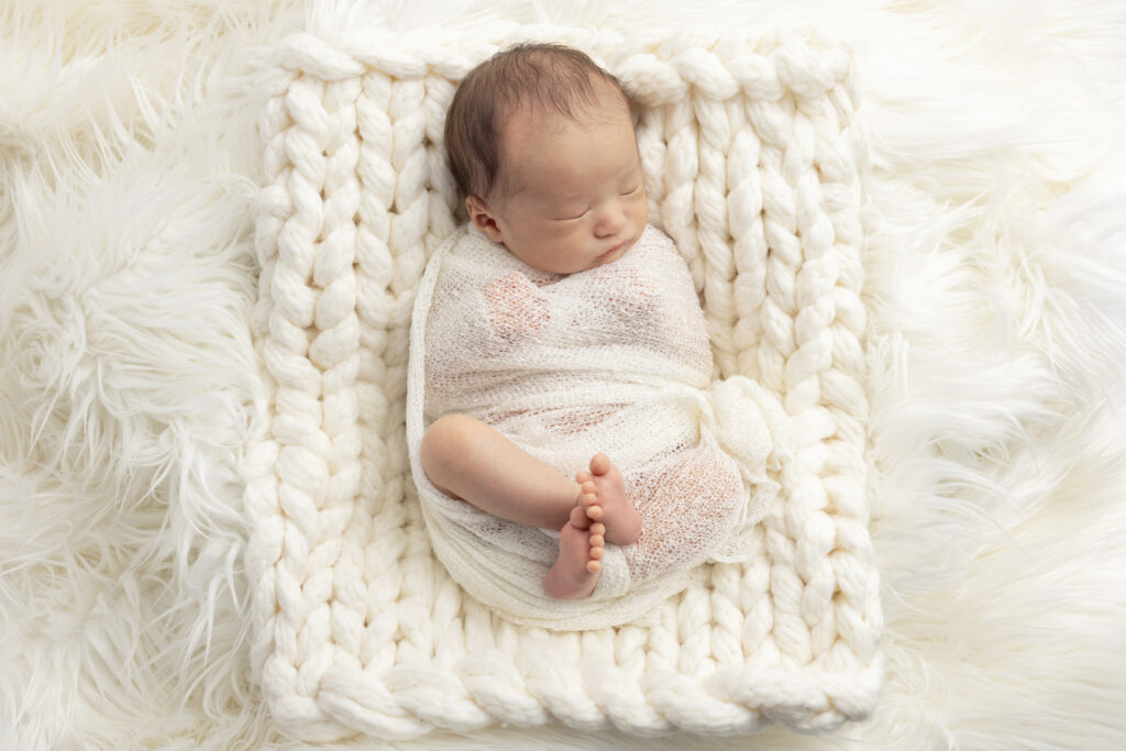 a newborn baby boy is lightly swaddled in ivory and lays on a chunky knit off white blanket placed on an ivory flokati; photographed by Karen Kahn of Looking Up Photography
