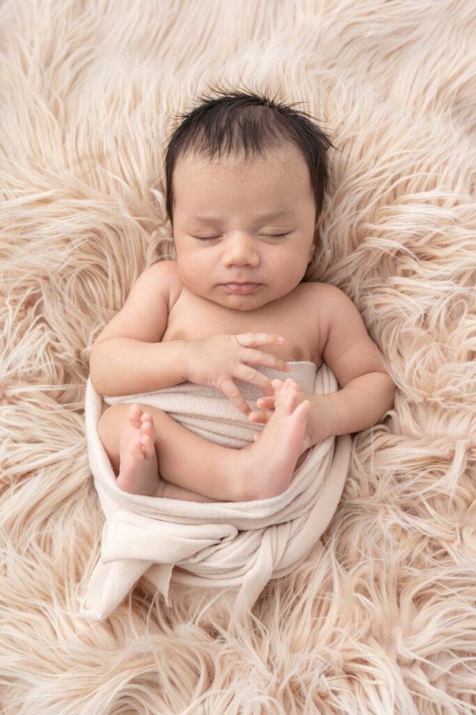 A newborn baby boy with dark hair sleeps peacefully in a relaxed pose on a creamy, light brown colored flokati, lightly wrapped in a felted textured swaddle.
