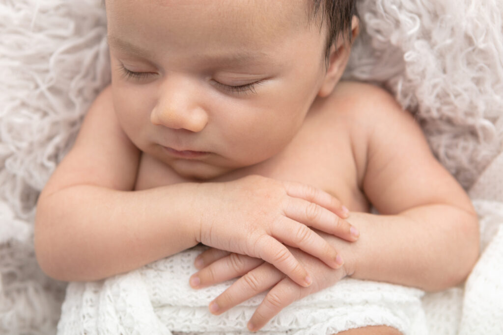 A newborn baby boy sleeps peacefully with his delicate hands folded across his stomach. He is lightly wrapped in a textured white blanket and lies on a light taupe colored flokati.
